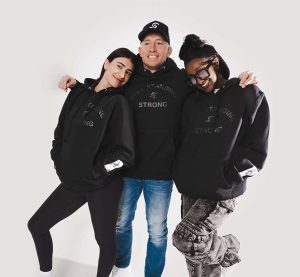 Georges St-Pierre pose avec deux femmes, tous vêtus de hoodies noirs arborant l'inscription "STILL STANDING STRONG" en ton sur ton. Ils affichent une posture détendue et complice, avec des sourires chaleureux. Georges porte également une casquette noire avec un logo blanc.